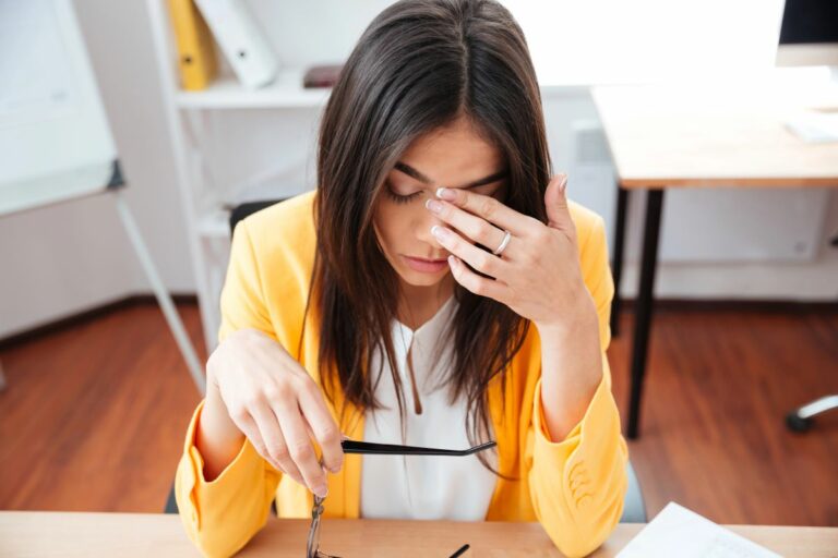 Woman sitting at her desk with glasses off finger on forehead